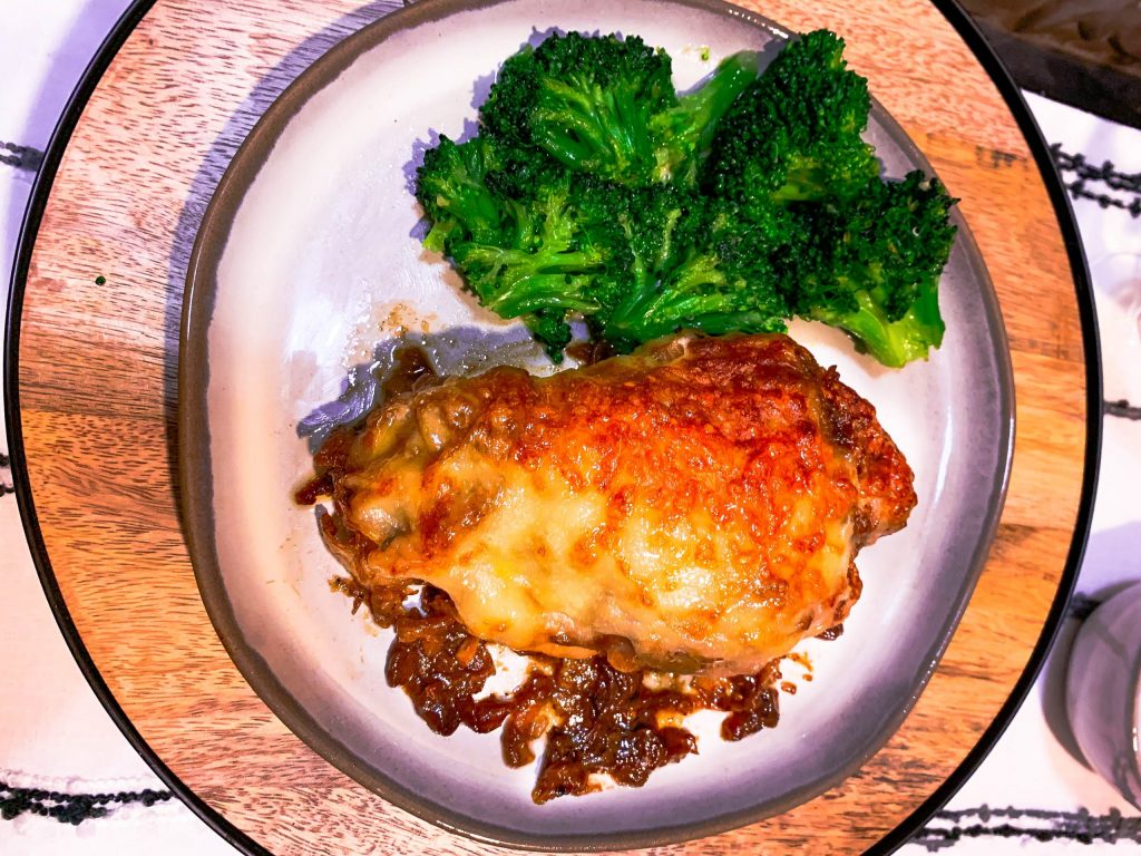 Roasted chicken breast covered in cartelized onions and Swiss cheese on a gray plate with steamed broccoli beside it.